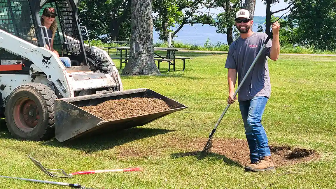 Two WoodChucks crew members filling and raking a ground stump with black dirt beside Lake Bemidji, one running the skid steer